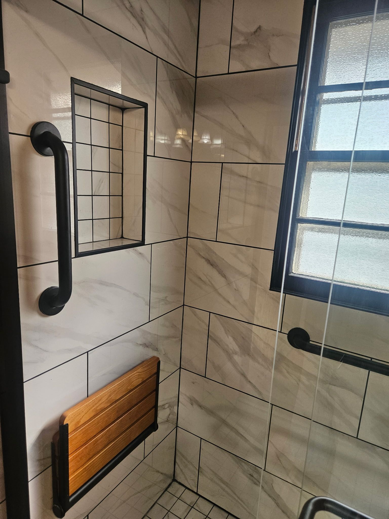 Accessible shower with grab bars, built-in seat, and recessed shelf. White marble-look tiles, black trim.