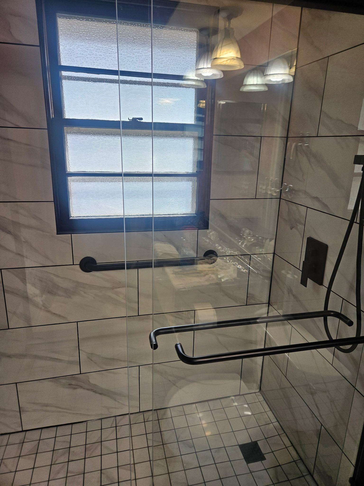 Shower with glass door, window, white tile, black fixtures, and a handle bar.
