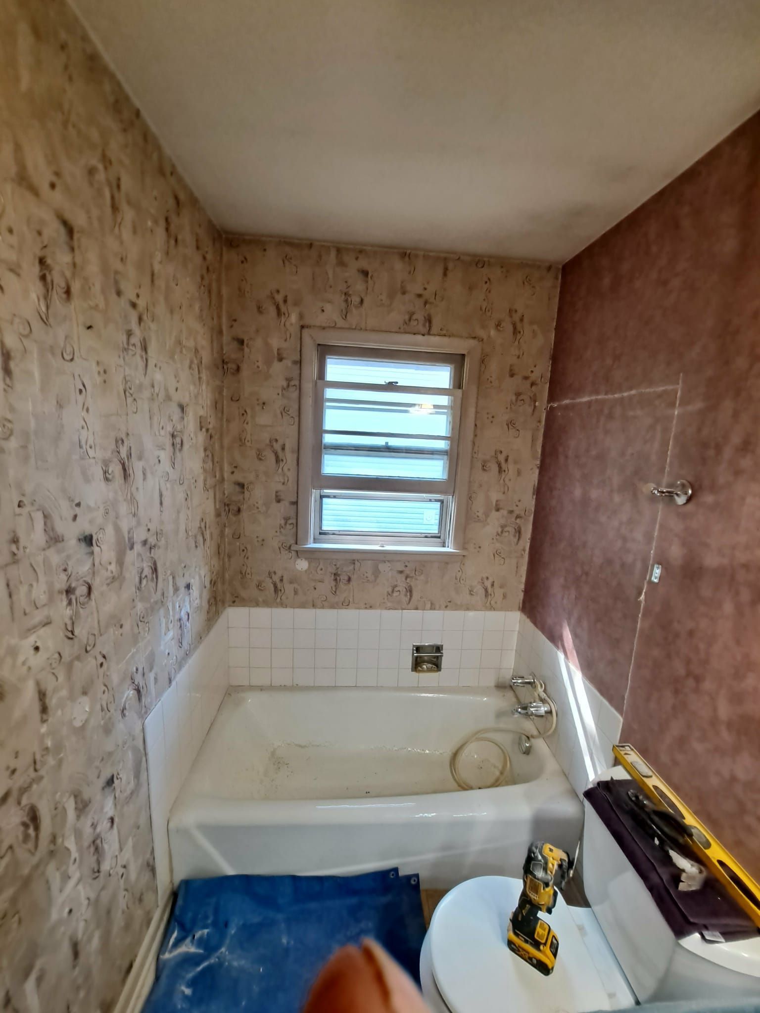Bathroom with wallpaper, tub, and window. Toilet and tools are visible.