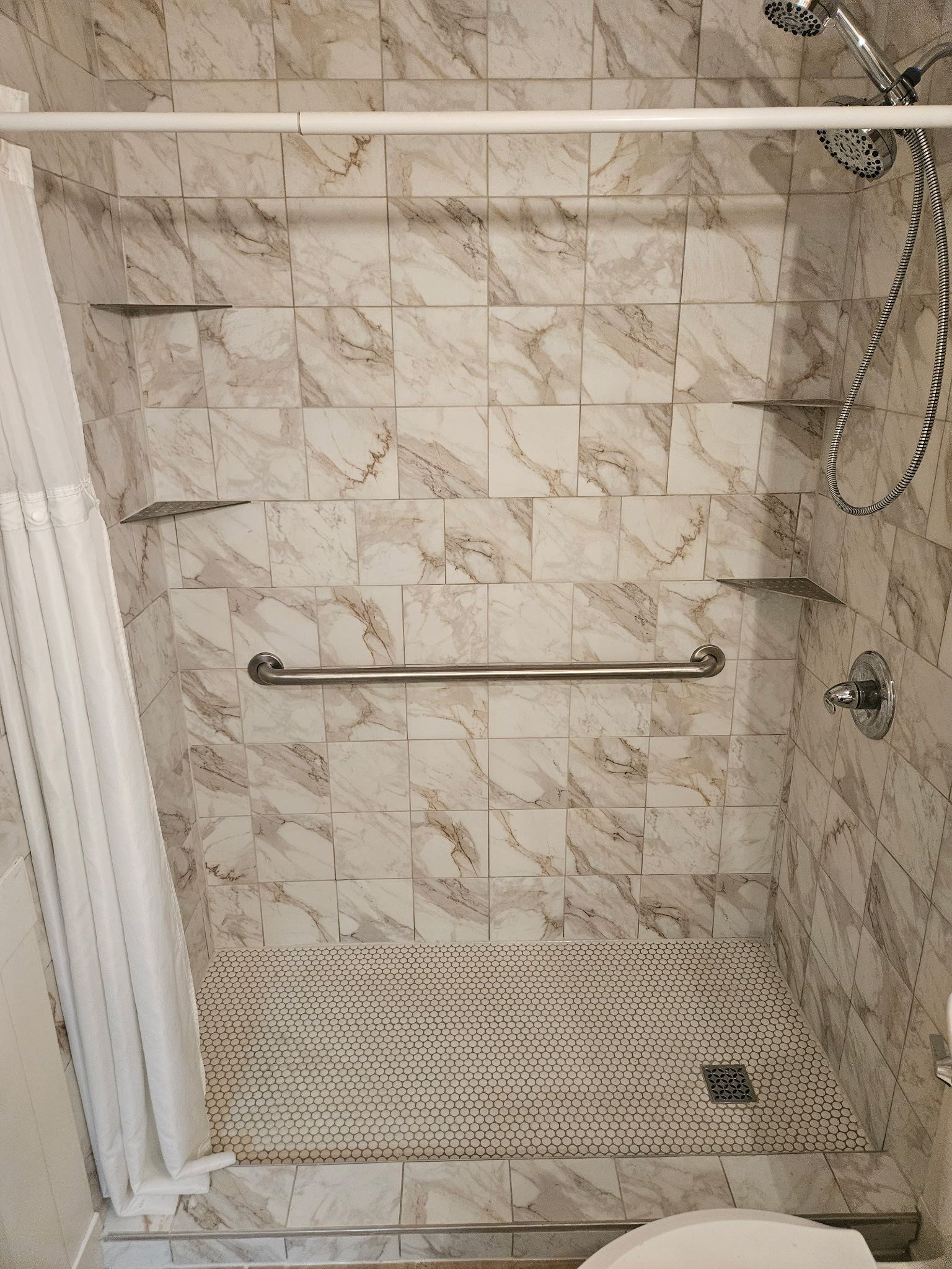 Shower stall with marble-patterned tile, grab bar, showerhead, and small shelves. White curtain on the left.