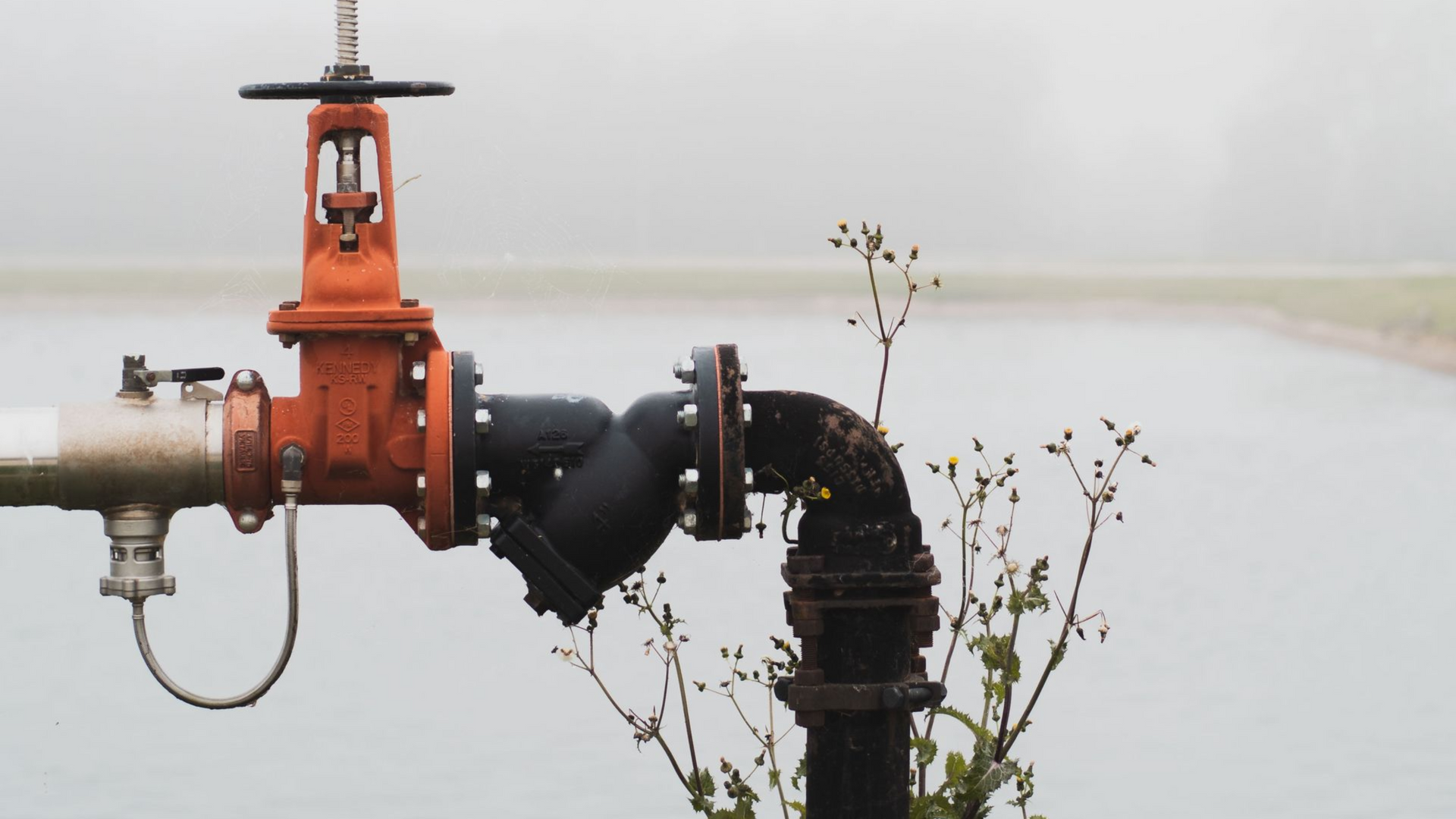 Orange water valve connected to black and metal pipes near water body.