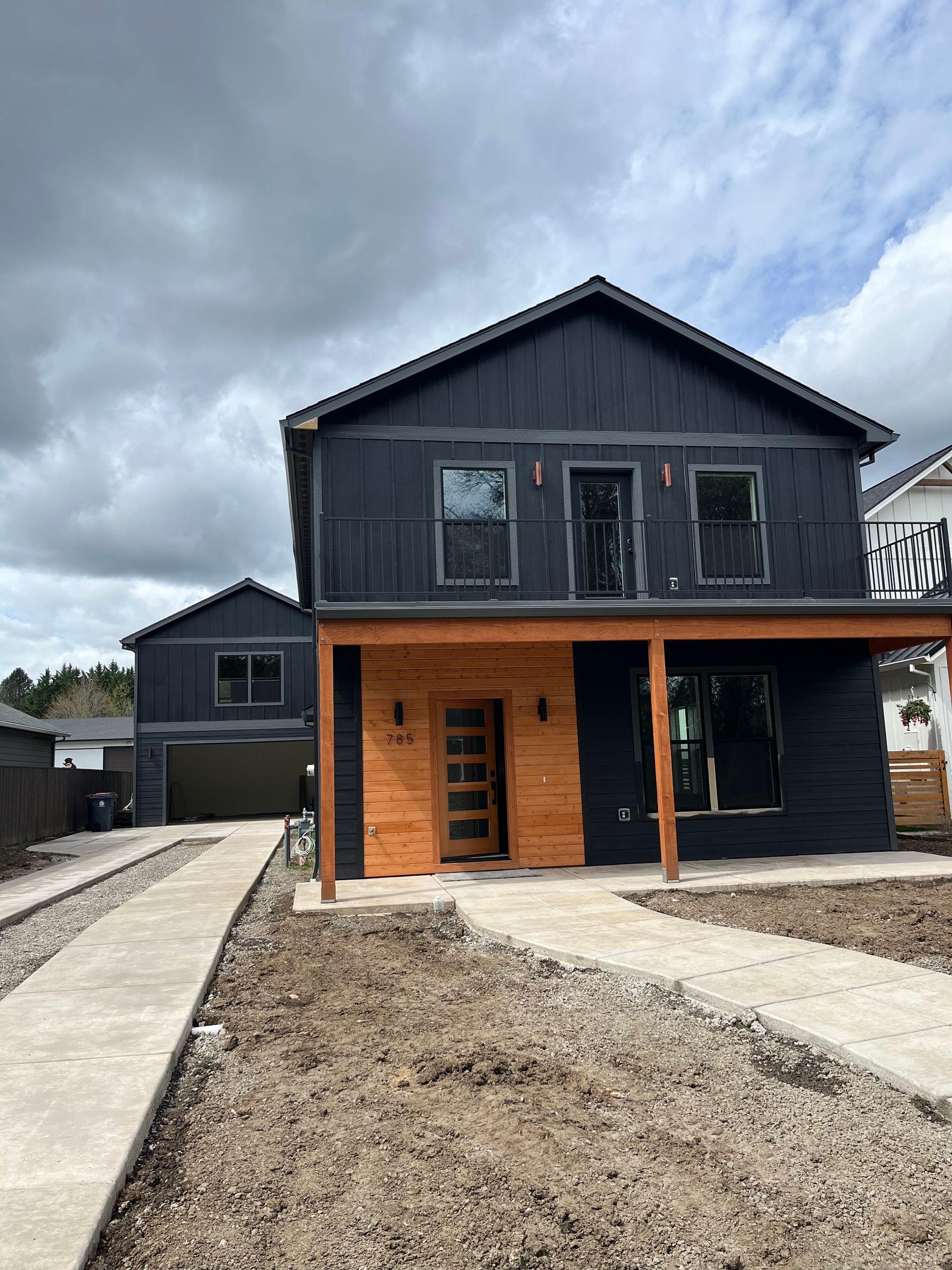 Modern two-story house with dark siding, wood porch accents, and a gravel driveway under cloudy skies
