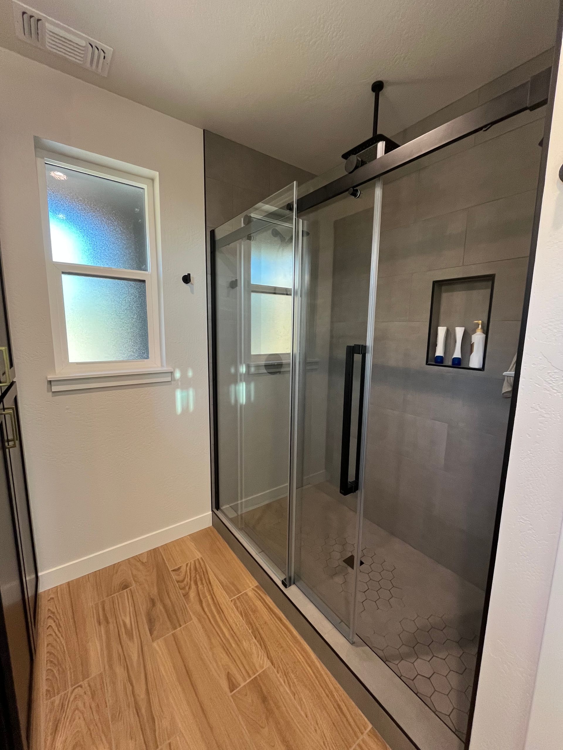 Modern bathroom with glass shower, gray tile, wood floor, and frosted window