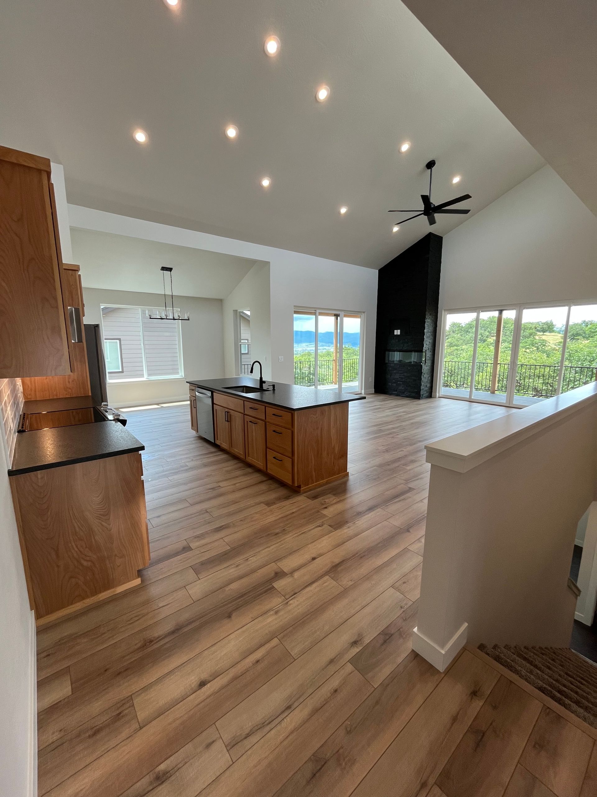 Open-plan kitchen and living area with wood floors, island, recessed lights, and large windows overlooking greenery
