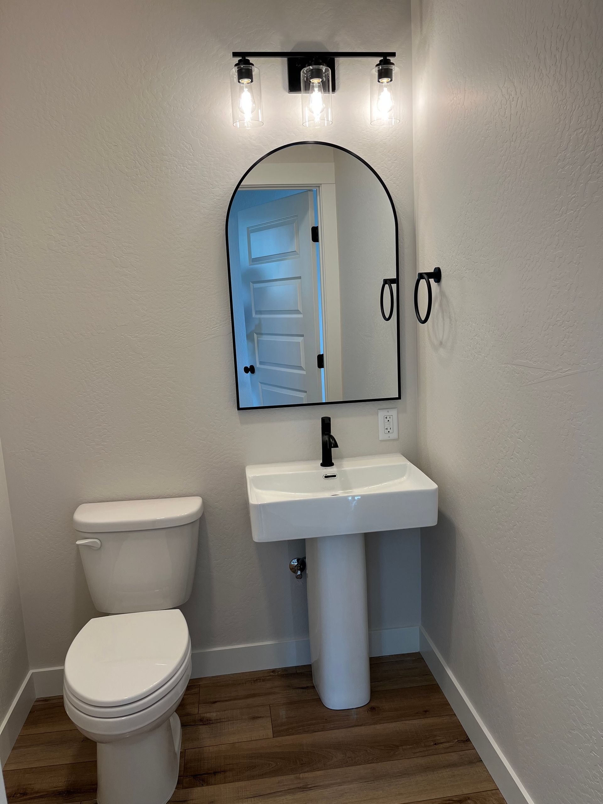 Small bathroom with toilet, pedestal sink, arched mirror, and wood-look flooring.
