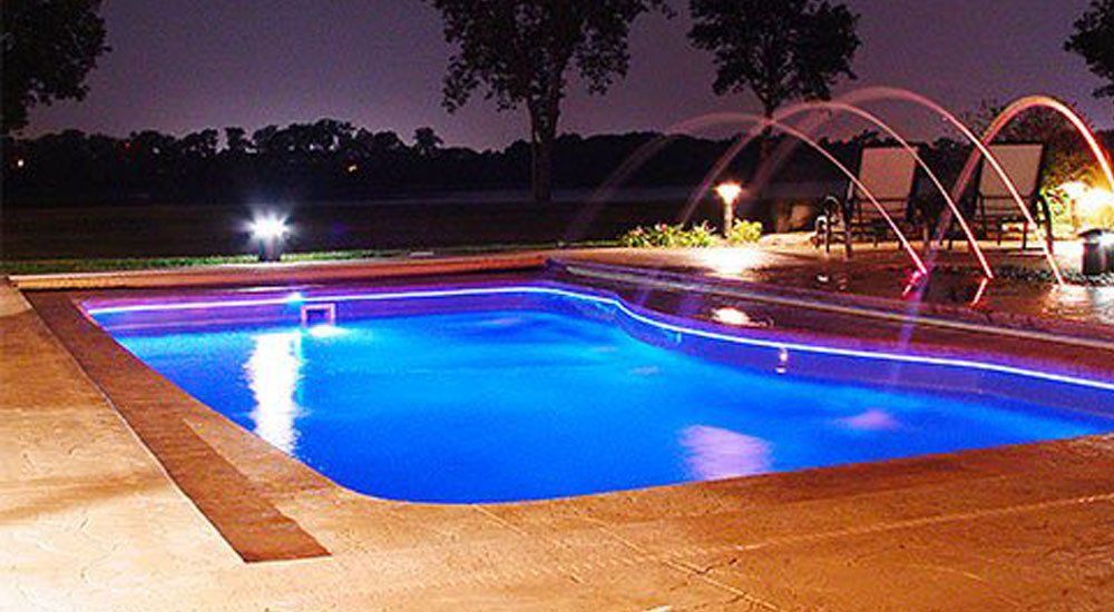 A large swimming pool is lit up at night with a fountain in the background.