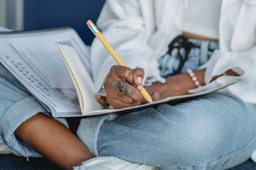 A woman is sitting on the floor writing in a notebook with a pencil.