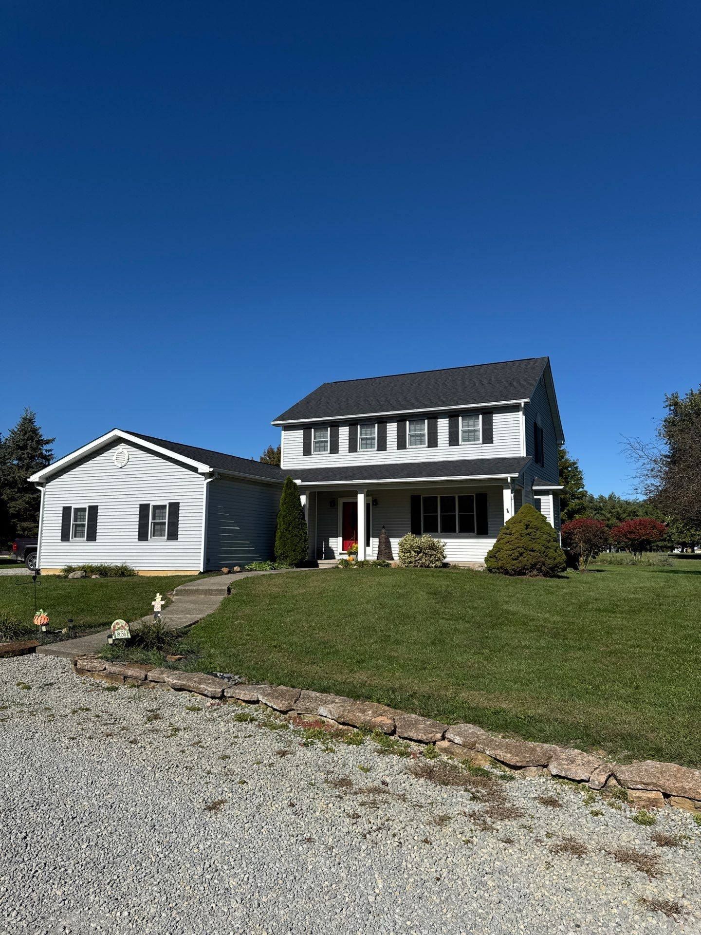 Two-story white house with attached garage on a sunny day, set on a grassy hill with a gravel driveway.