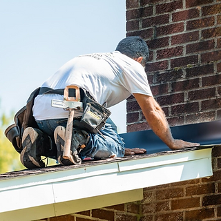 Worker kneeling on a roof edge, installing shingles beside a brick chimney