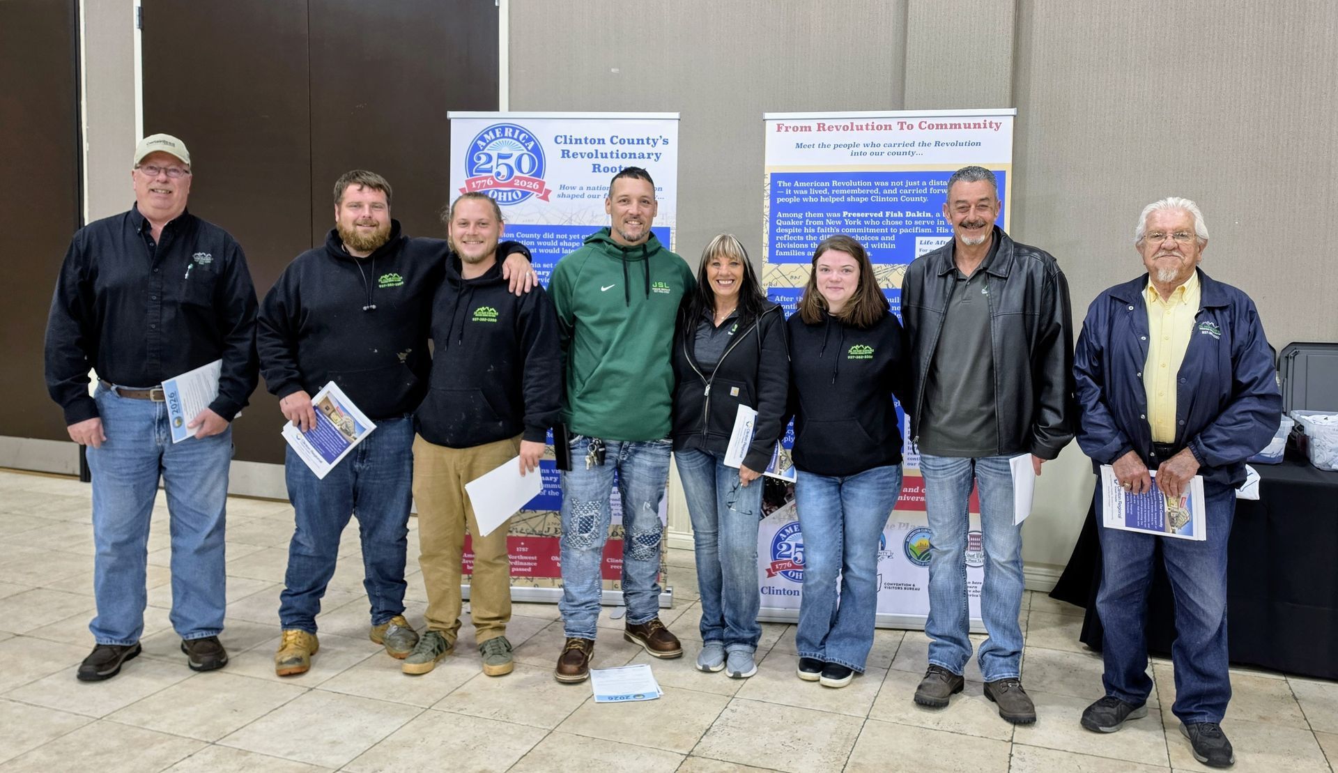 Seven people standing indoors in winter coats, posing in front of banners on a concrete floor.