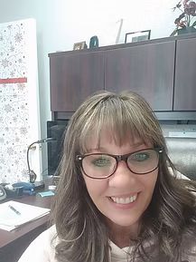 Smiling woman with glasses in an office, seated near a desk and dark cabinets.