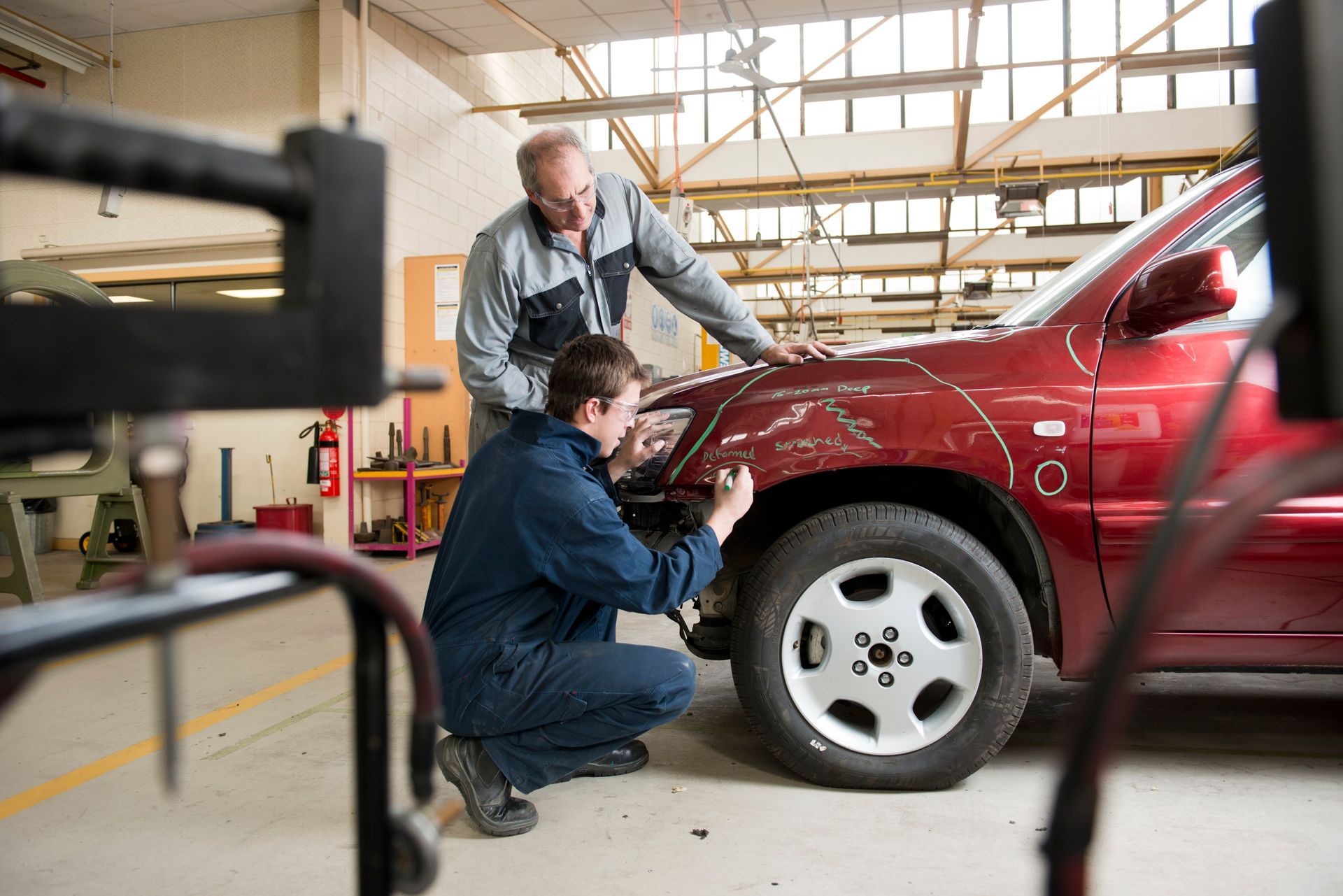 Two mechanics inspecting the front wheel of a red car in a garage