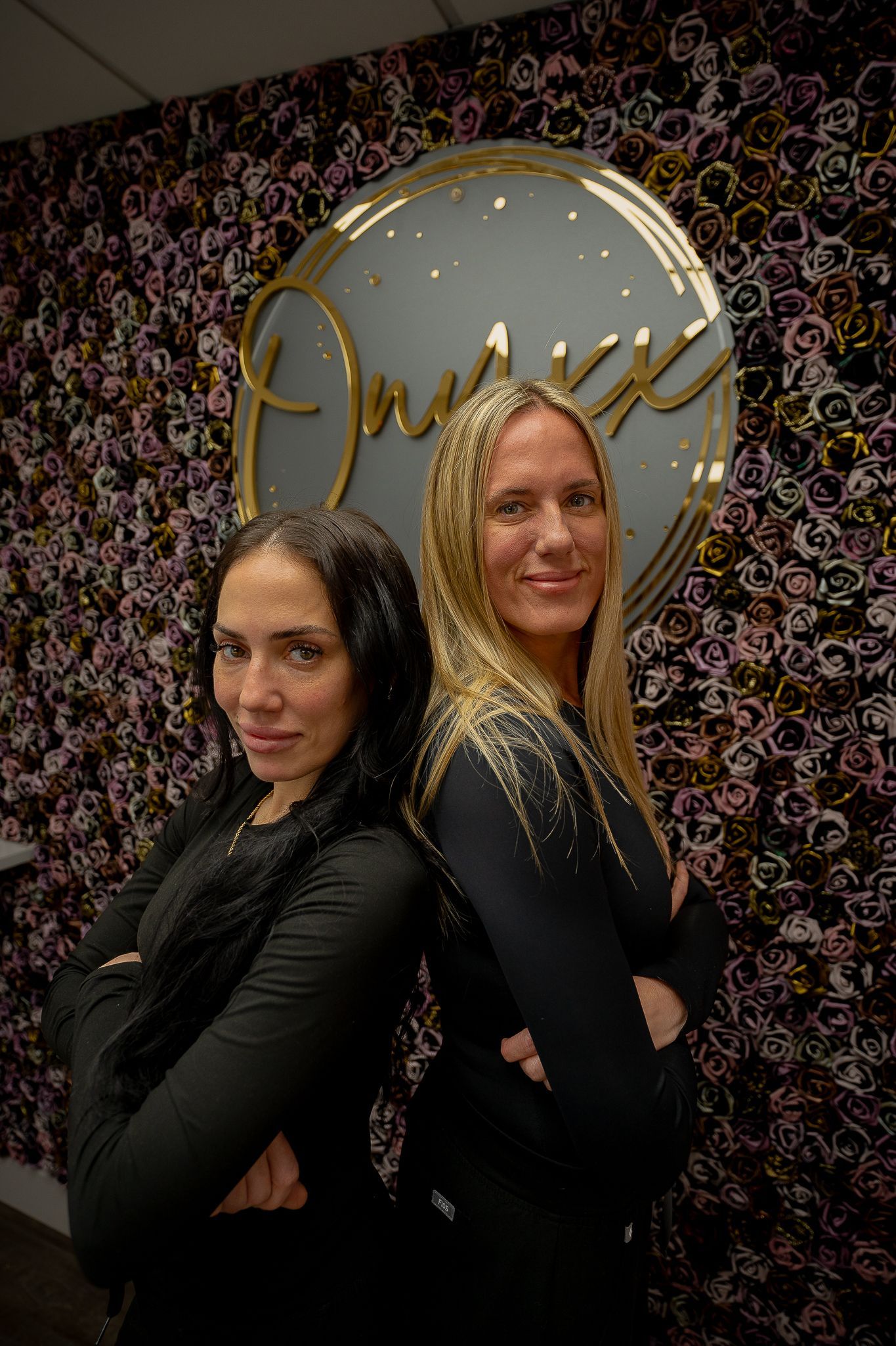 Two women are standing back to back in front of a floral wall.