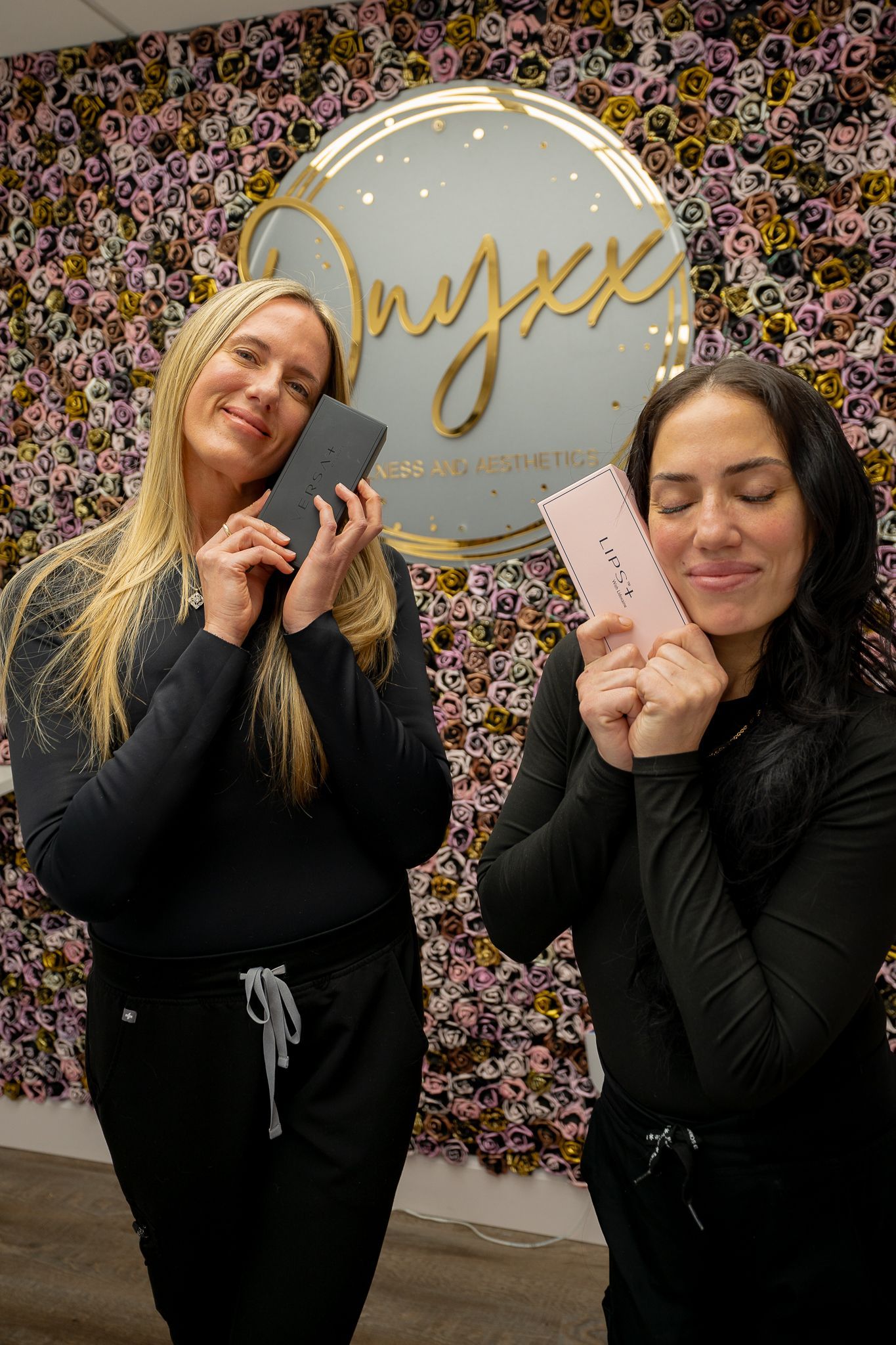 Two women are standing next to each other in front of a flower wall holding cell phones.