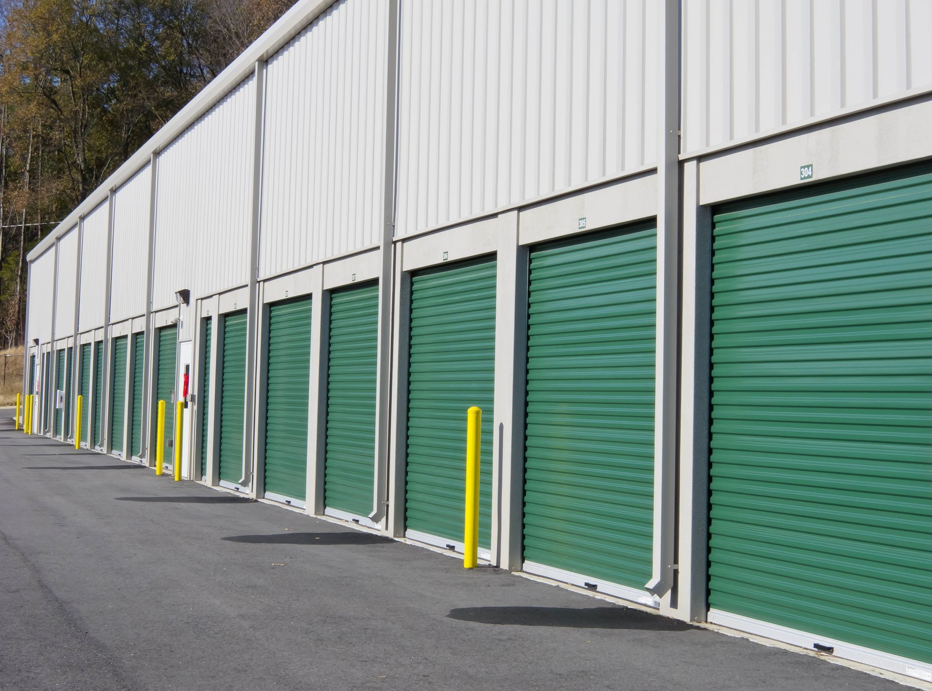 Storage units in a row; green doors, white building, asphalt road, yellow posts, outdoors.