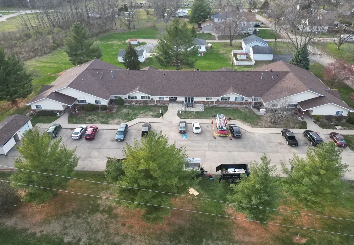 Aerial view of a long brown-roofed building with parked cars and trees in front.