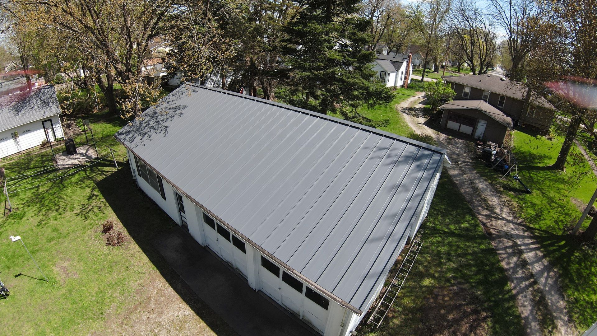 Aerial view of a long white building with a gray roof, surrounded by trees and a yard