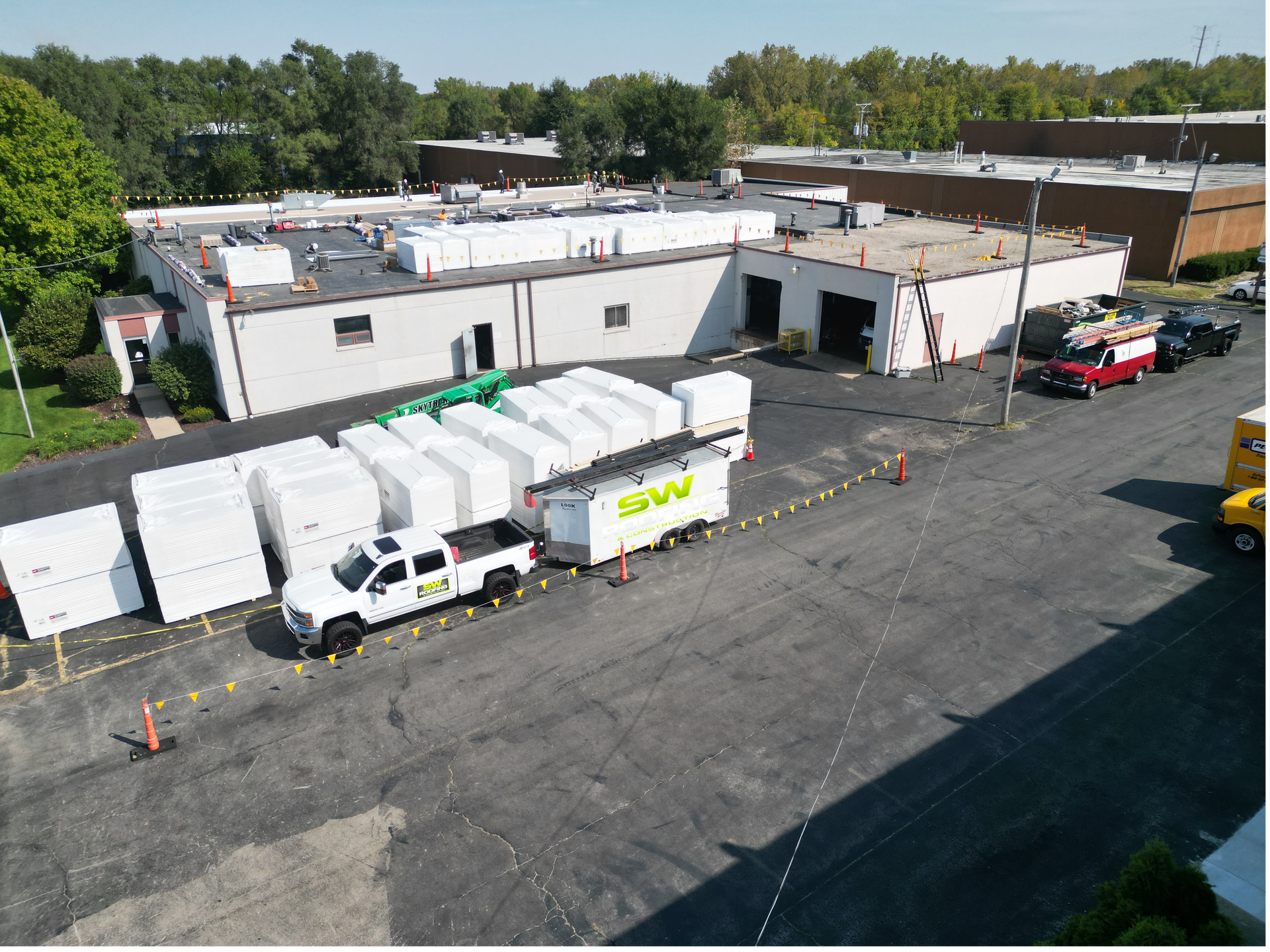 Aerial view of a white industrial building with trucks, trailers, and a parking lot under construction.