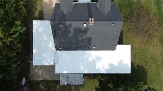 Aerial view of a house with a dark gray roof and white deck, surrounded by trees and grass.