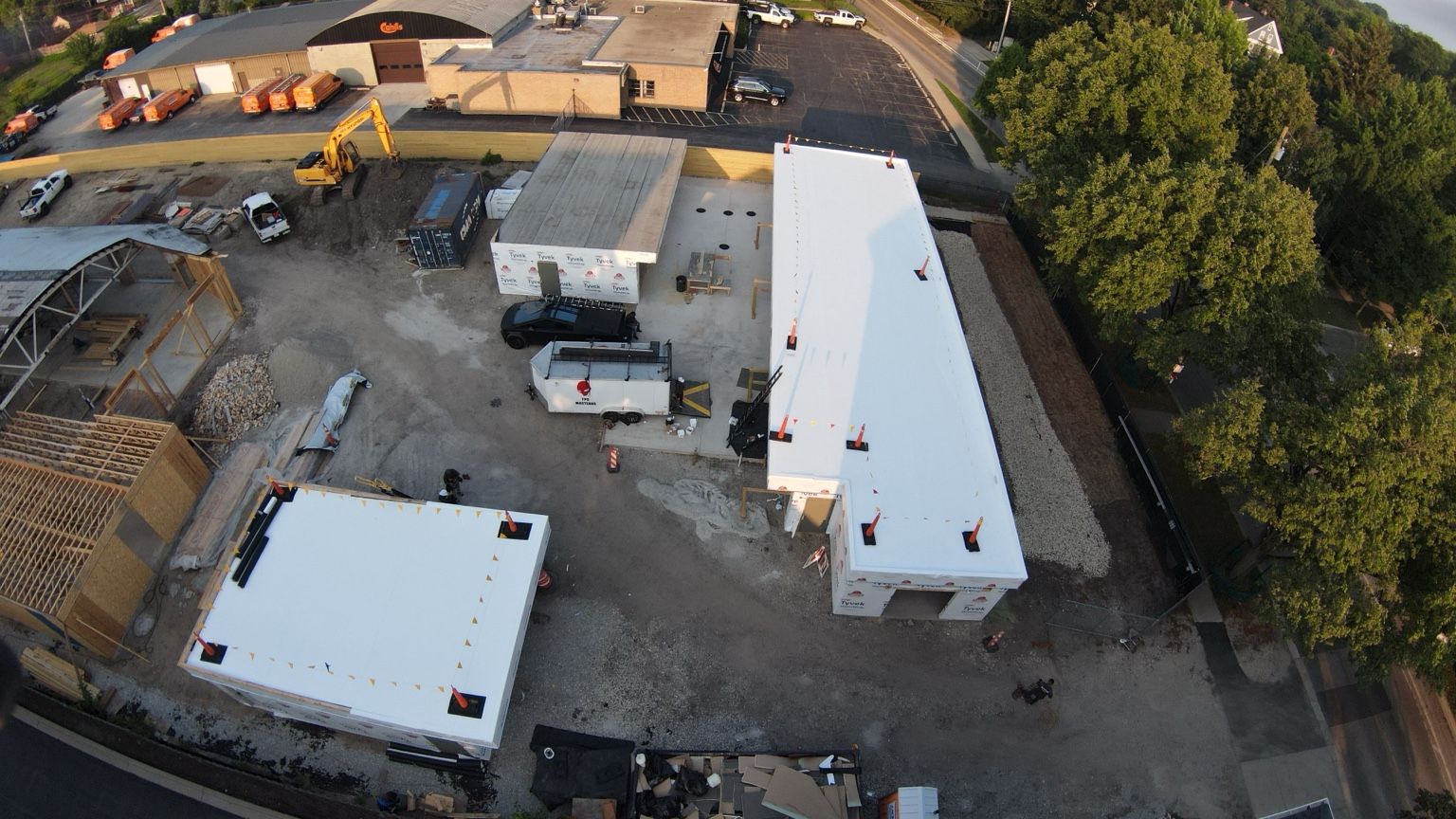 Aerial view of a construction site with white-roofed buildings, trucks, equipment, and trees.