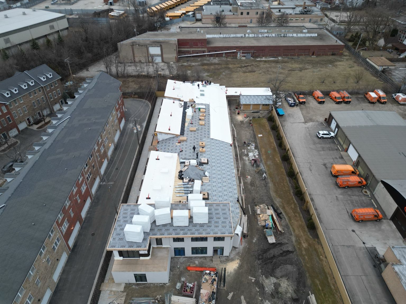 Aerial view of a long white-roofed building beside an industrial yard with parked orange trucks.