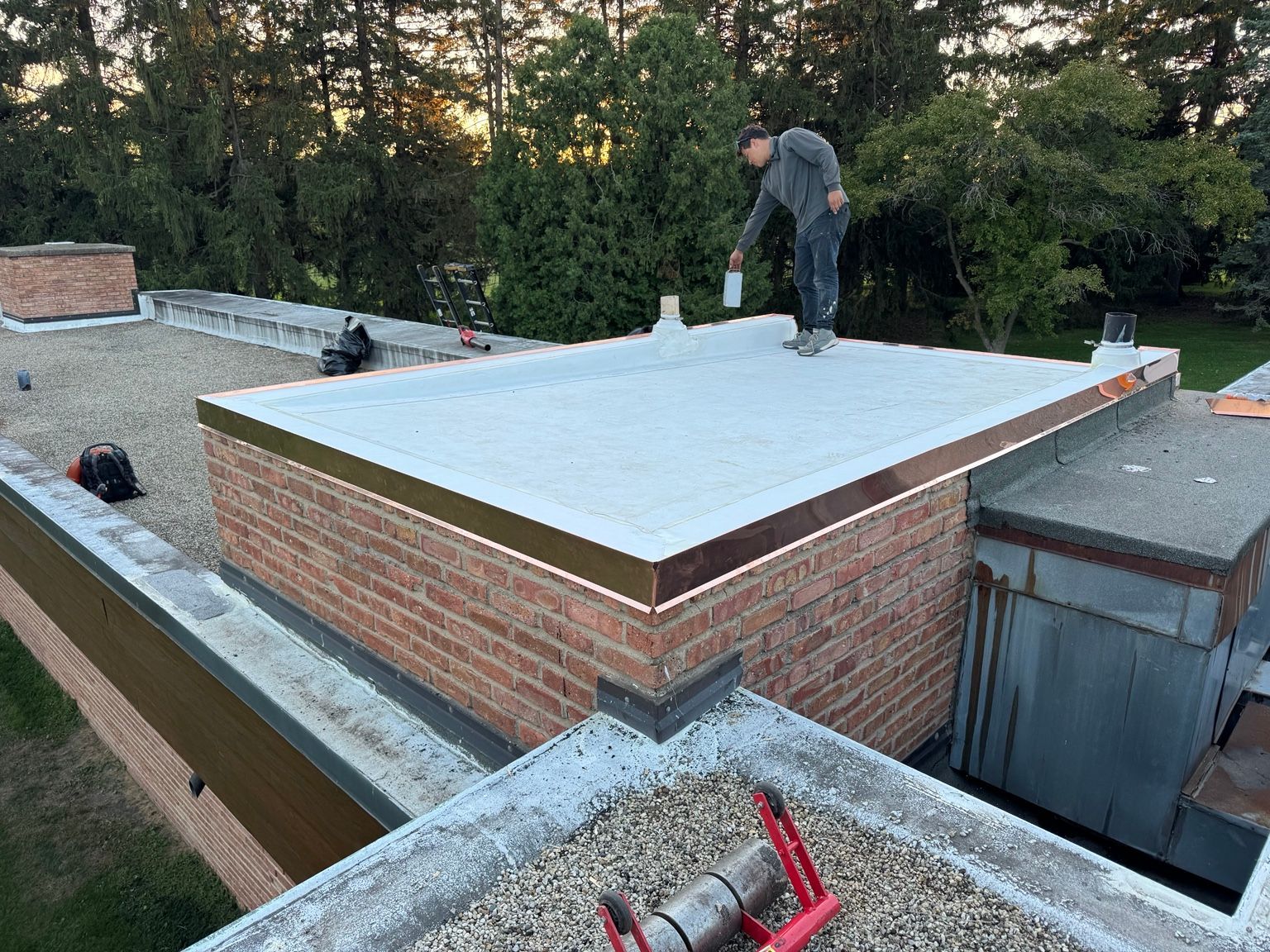 Worker on a flat rooftop applying a white roof membrane beside brick chimneys and trees.