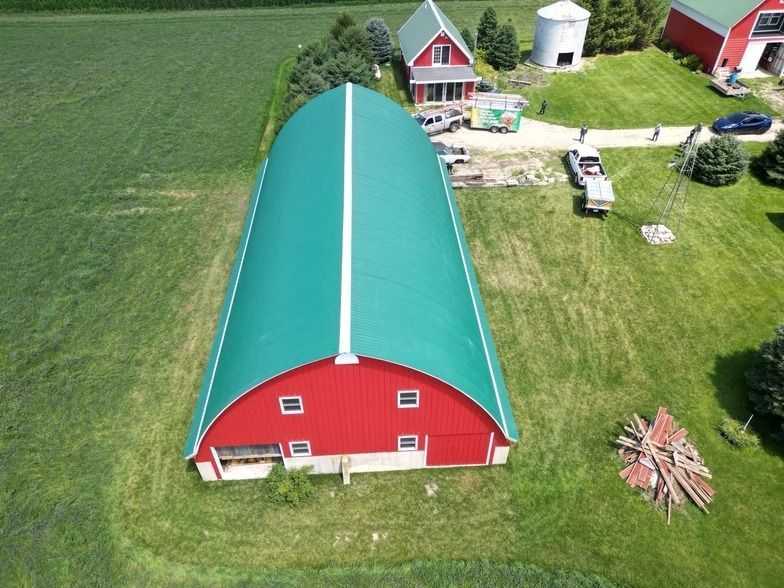 Aerial view of a red barn with a green roof beside a farmhouse and outbuildings on a grassy farm.