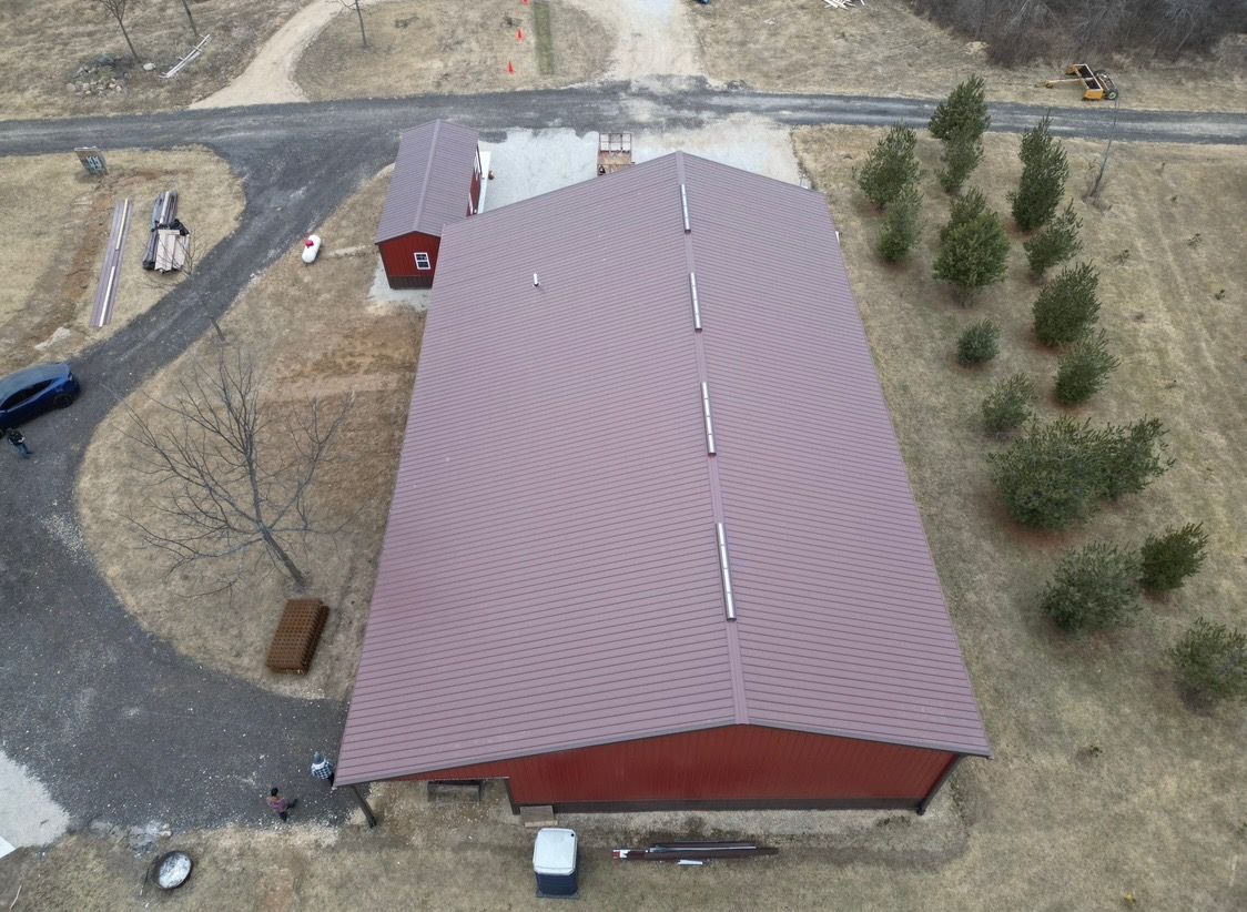 Aerial view of a large red barn with a metal roof, gravel driveway, and trees in a rural area