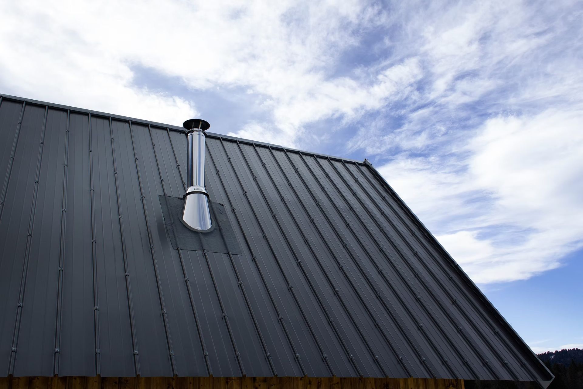 Dark metal roof with a silver chimney vent against a cloudy blue sky