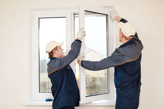 Two workers installing a white window frame in a bright room