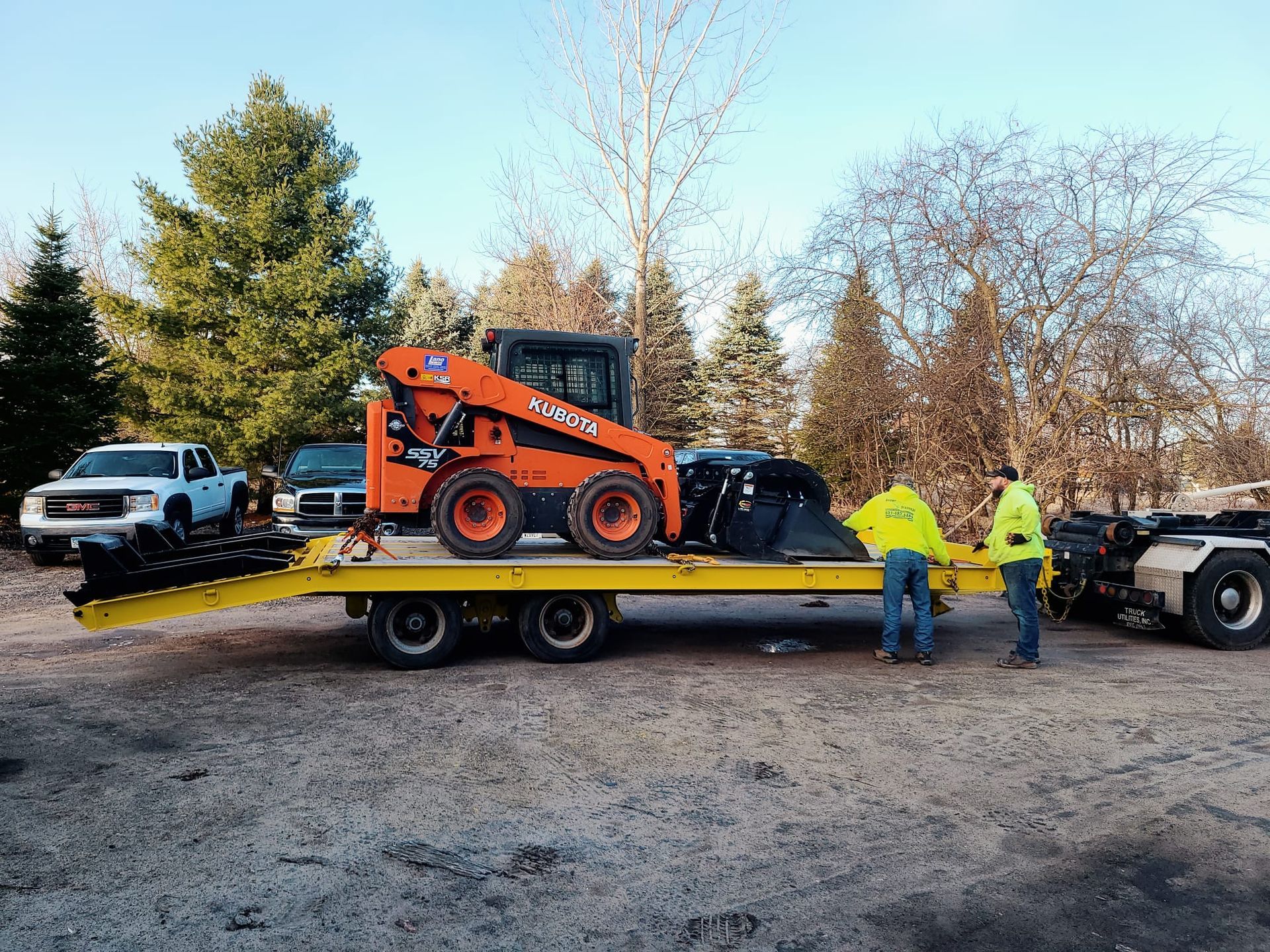 An orange skid steer on a yellow trailer being loaded by two people in safety vests.