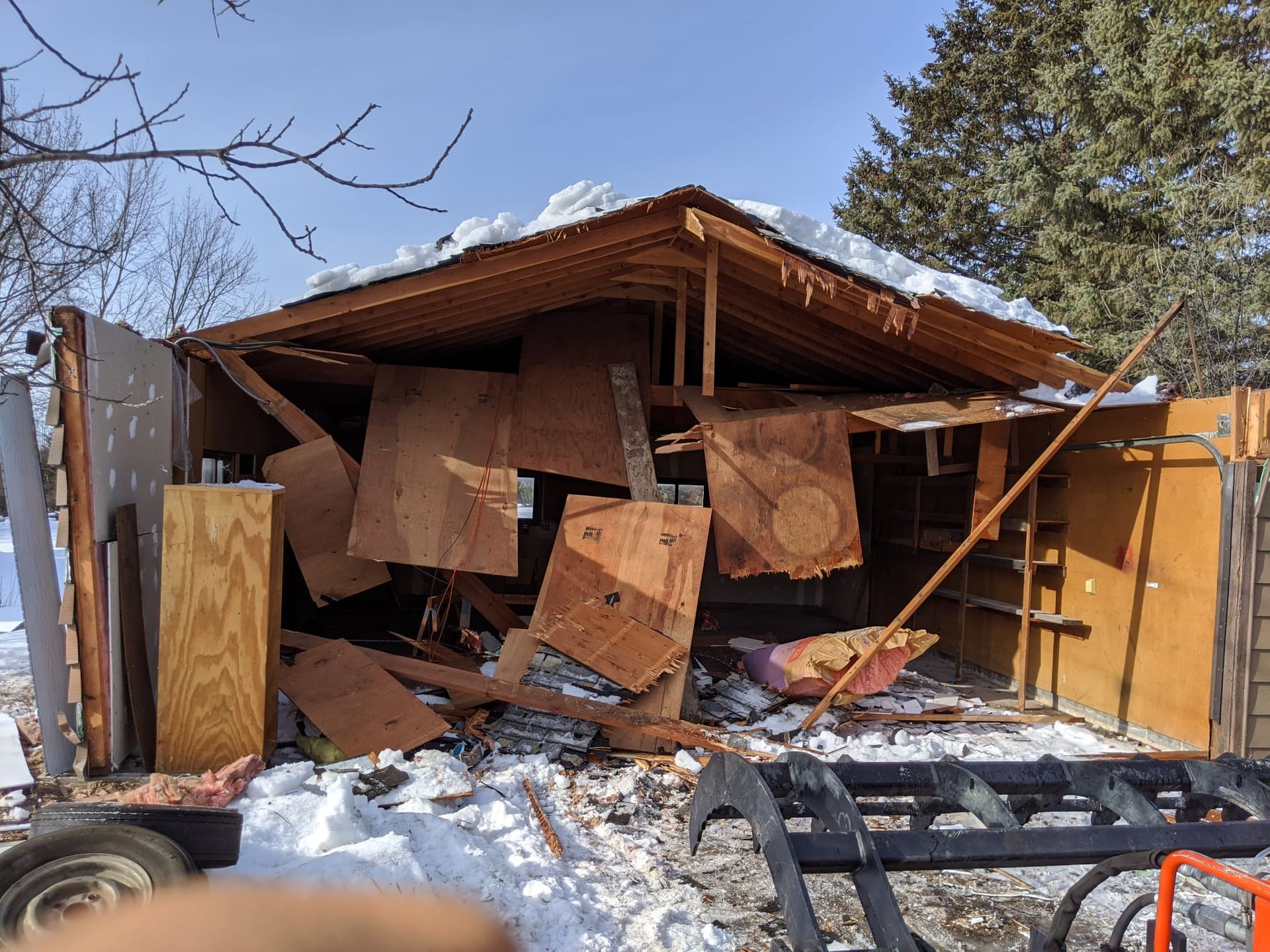 A partially collapsed wooden shed, debris on snow, under blue sky.