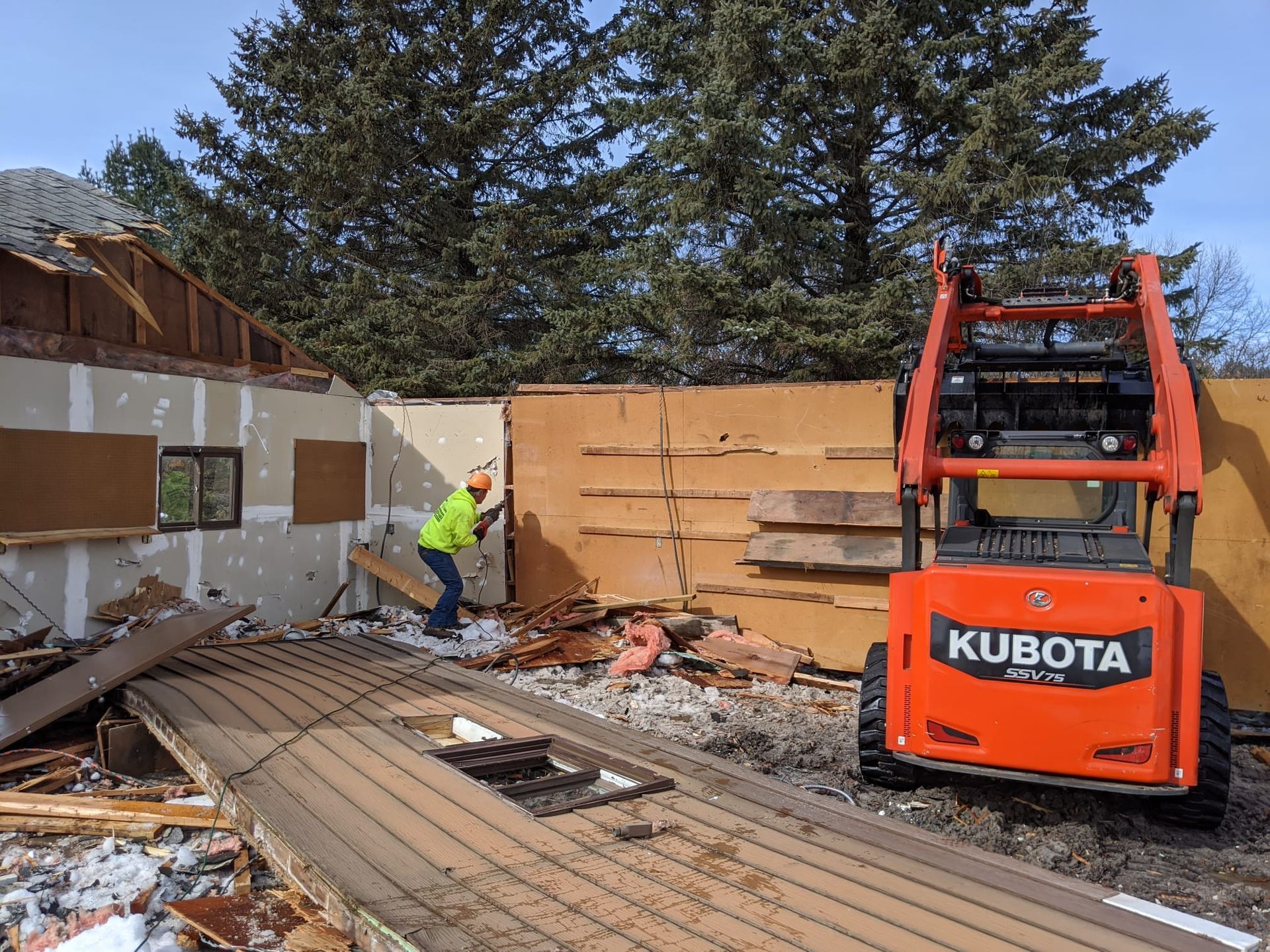 Demolition site: worker in safety vest using a tool. Orange Kubota machine stands nearby. Building partially torn down.