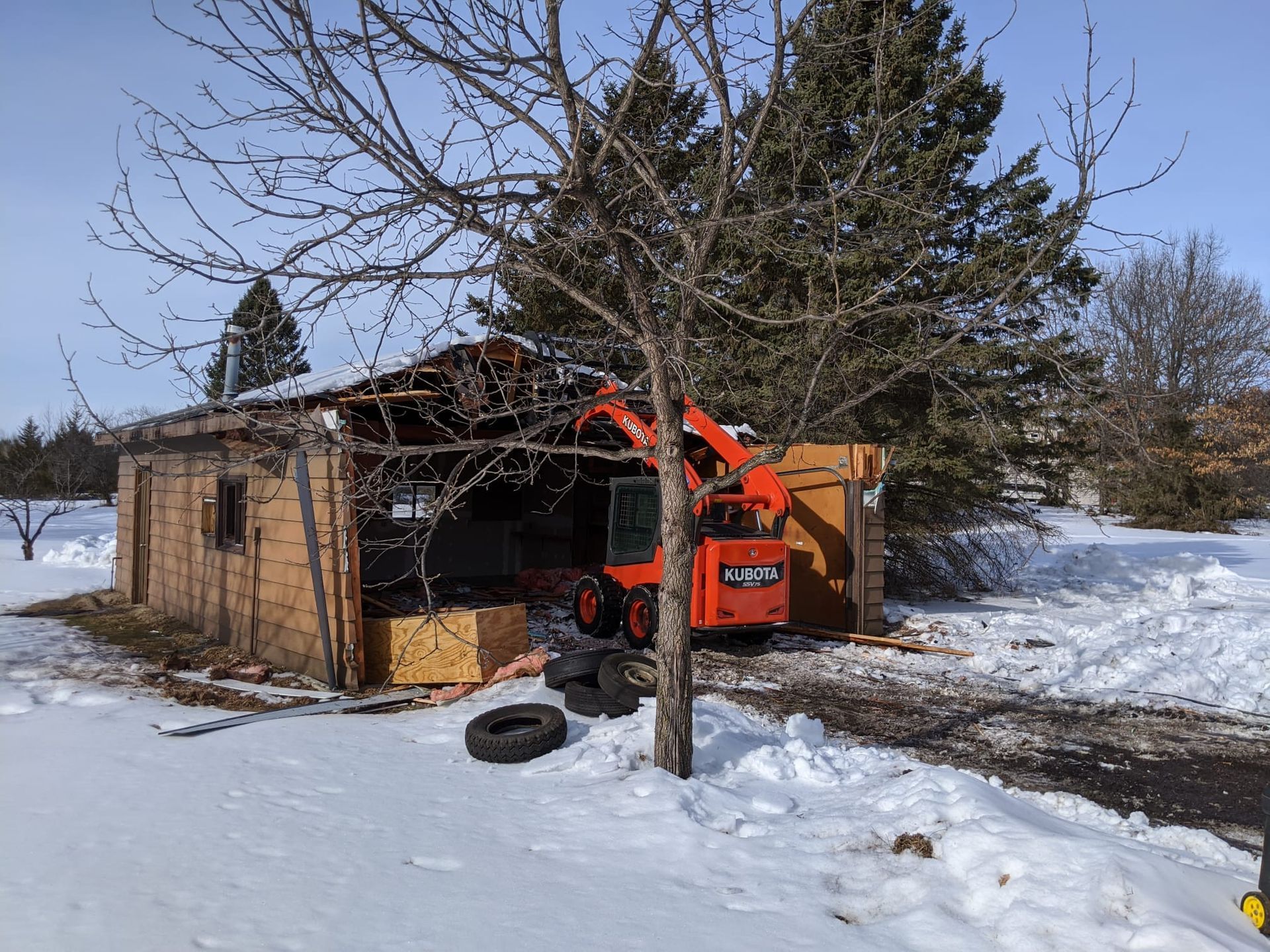A small excavator demolishing a wooden shed in a snowy outdoor setting.