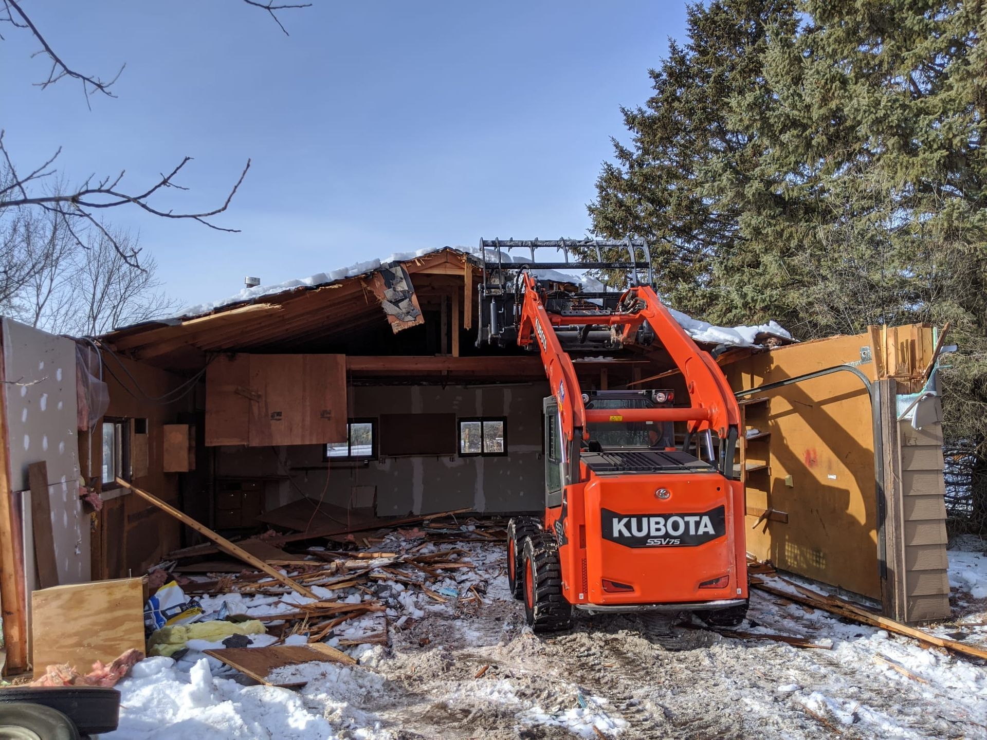 Orange Kubota skid steer demolishing a building with roof damage, snow on ground, clear sky.