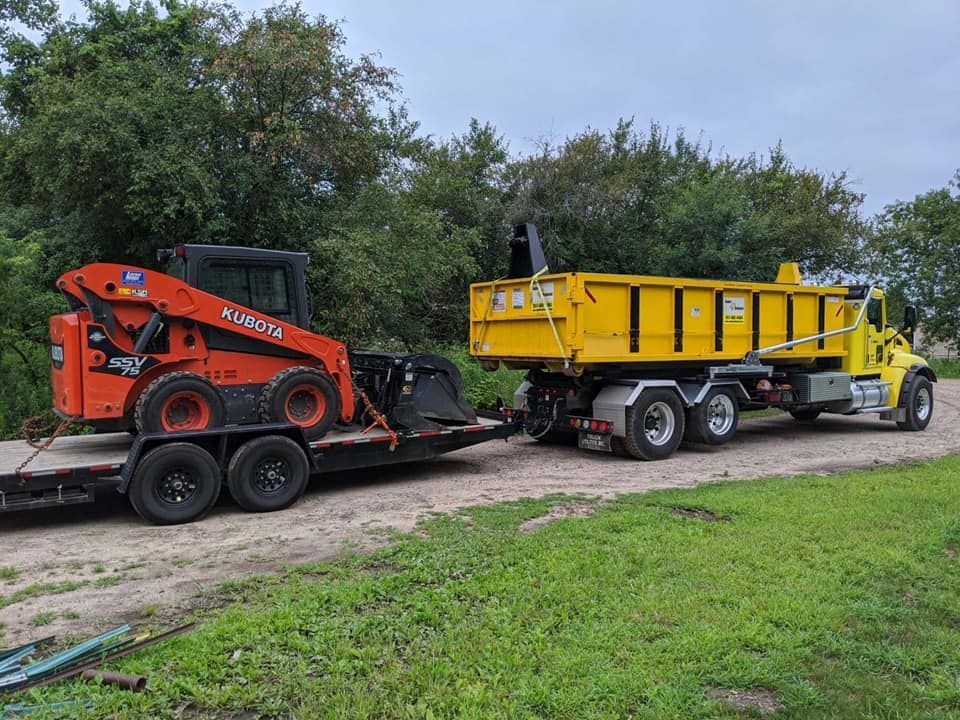 Orange skid steer on a trailer, towed by a yellow dump truck on a dirt road, trees in the background.
