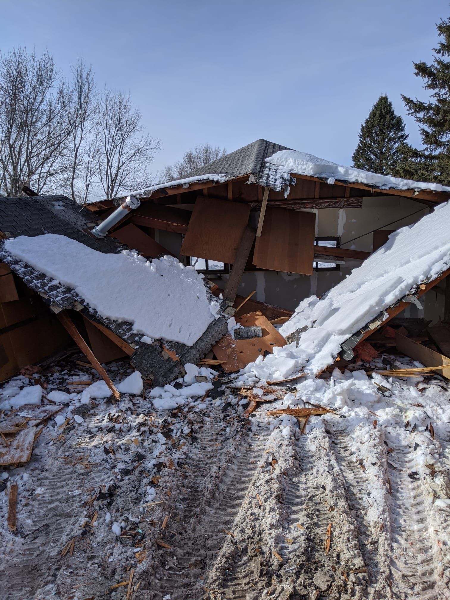 Partially demolished building with snow on the roof and debris. Winter setting, tire tracks in the foreground.