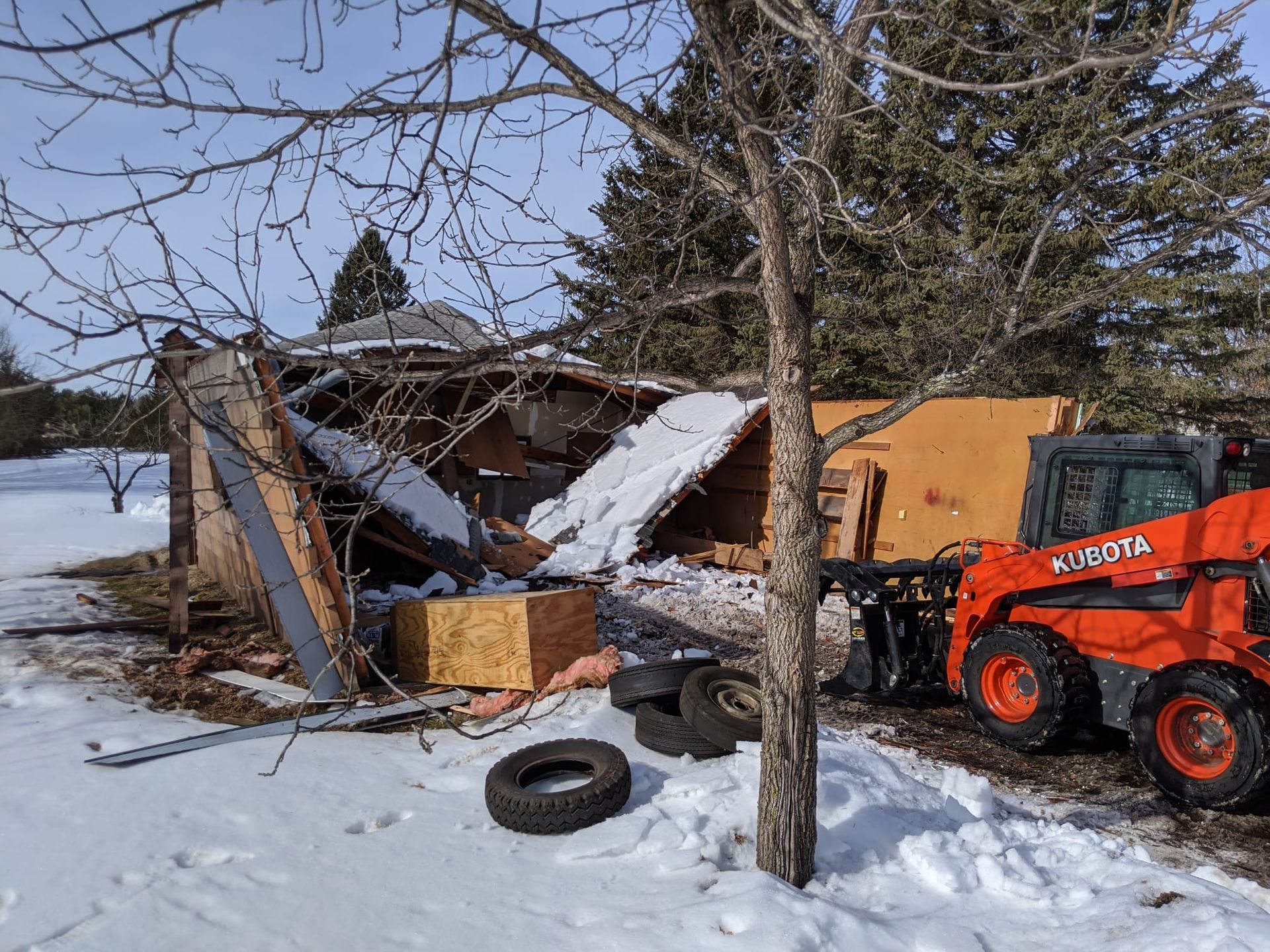 A partially demolished wooden shed in a snowy yard, with an orange Kubota skid steer nearby.