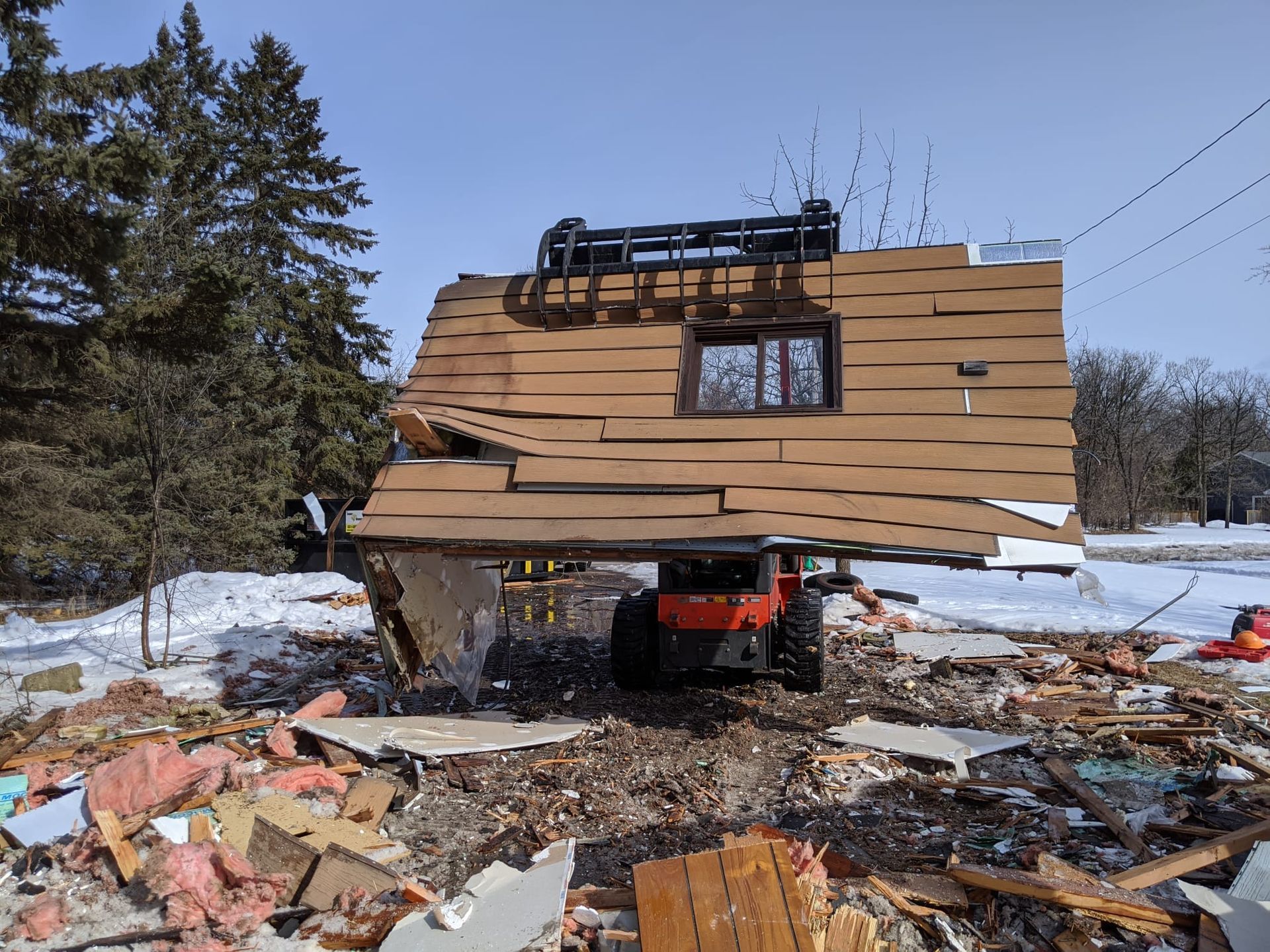 A small building's roof section is held by a forklift during demolition on a snowy day.
