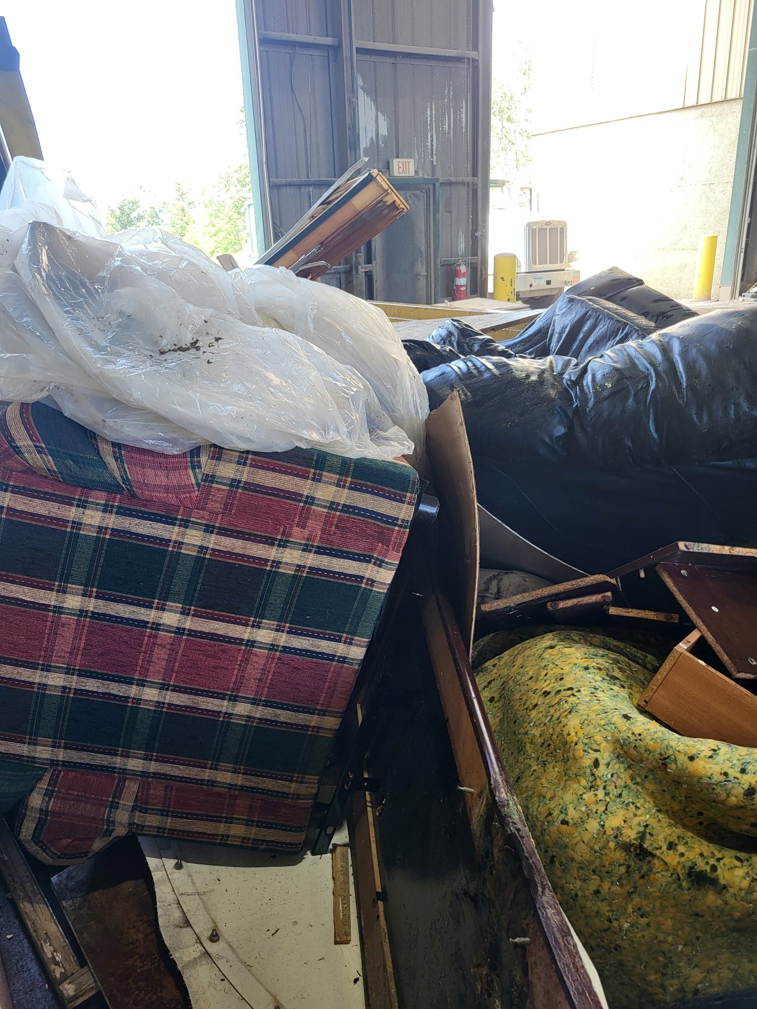 Pile of discarded furniture and debris inside a warehouse; plaid fabric, black bags, and wood are visible.