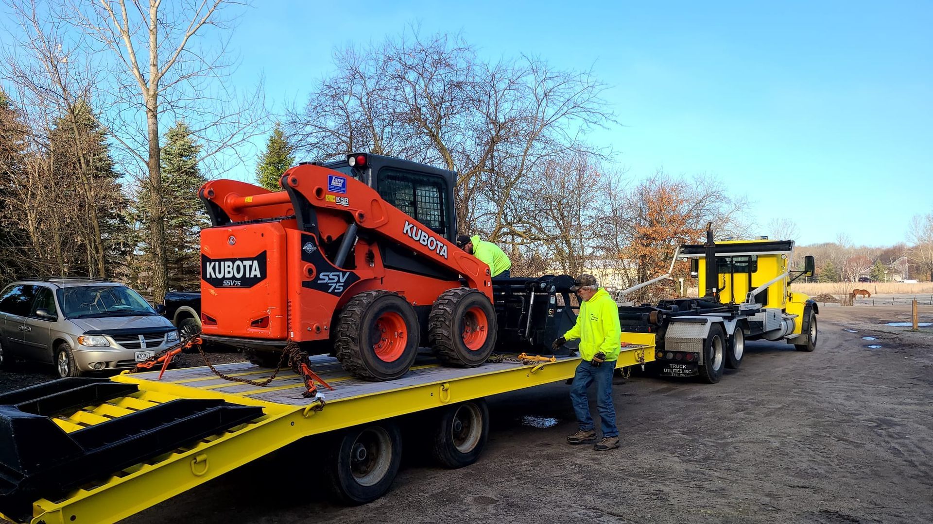 Orange skid steer loader secured on a yellow flatbed trailer, being loaded by two people next to a yellow semi-truck on a gravel lot.