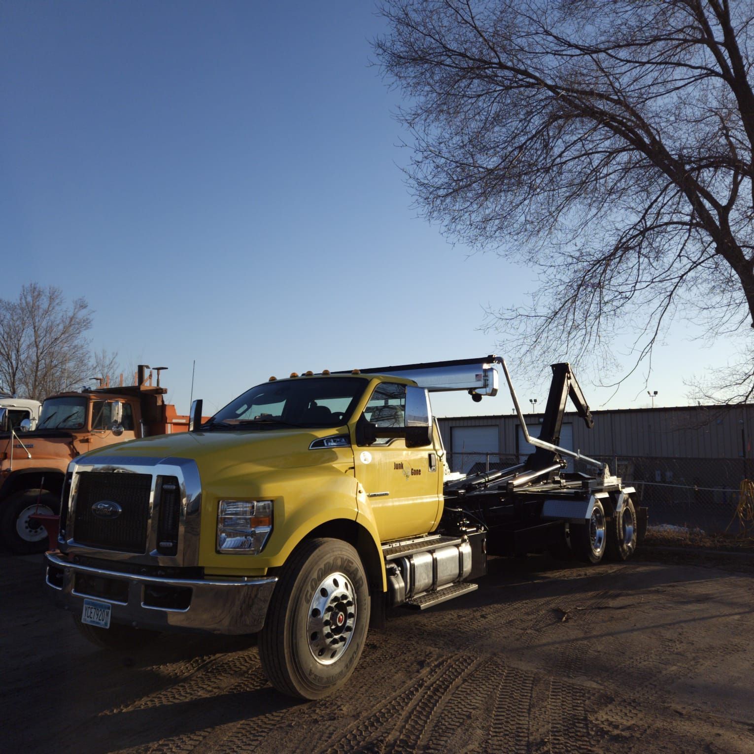 Yellow roll-off truck parked on dirt lot with another truck in the background, under a blue sky.