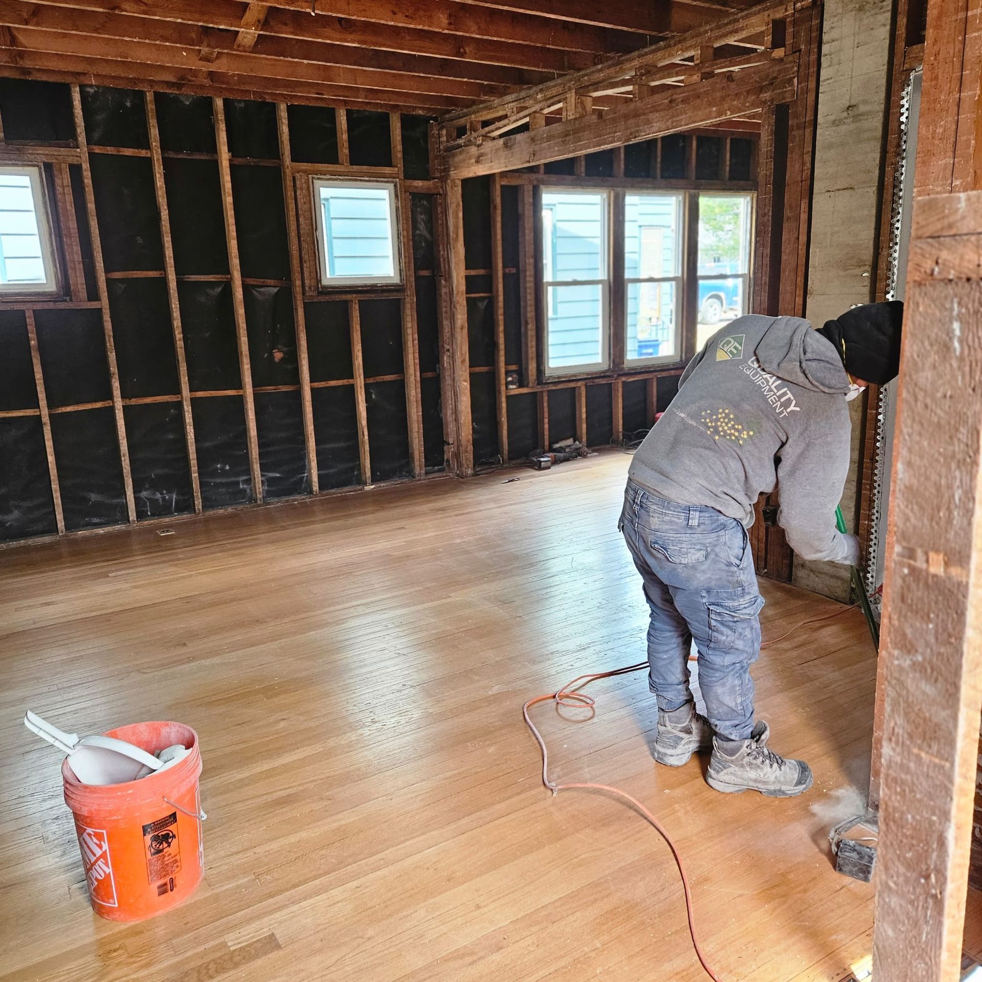 A person sanding a wooden floor in a room under construction. Orange bucket and electrical cord in view.
