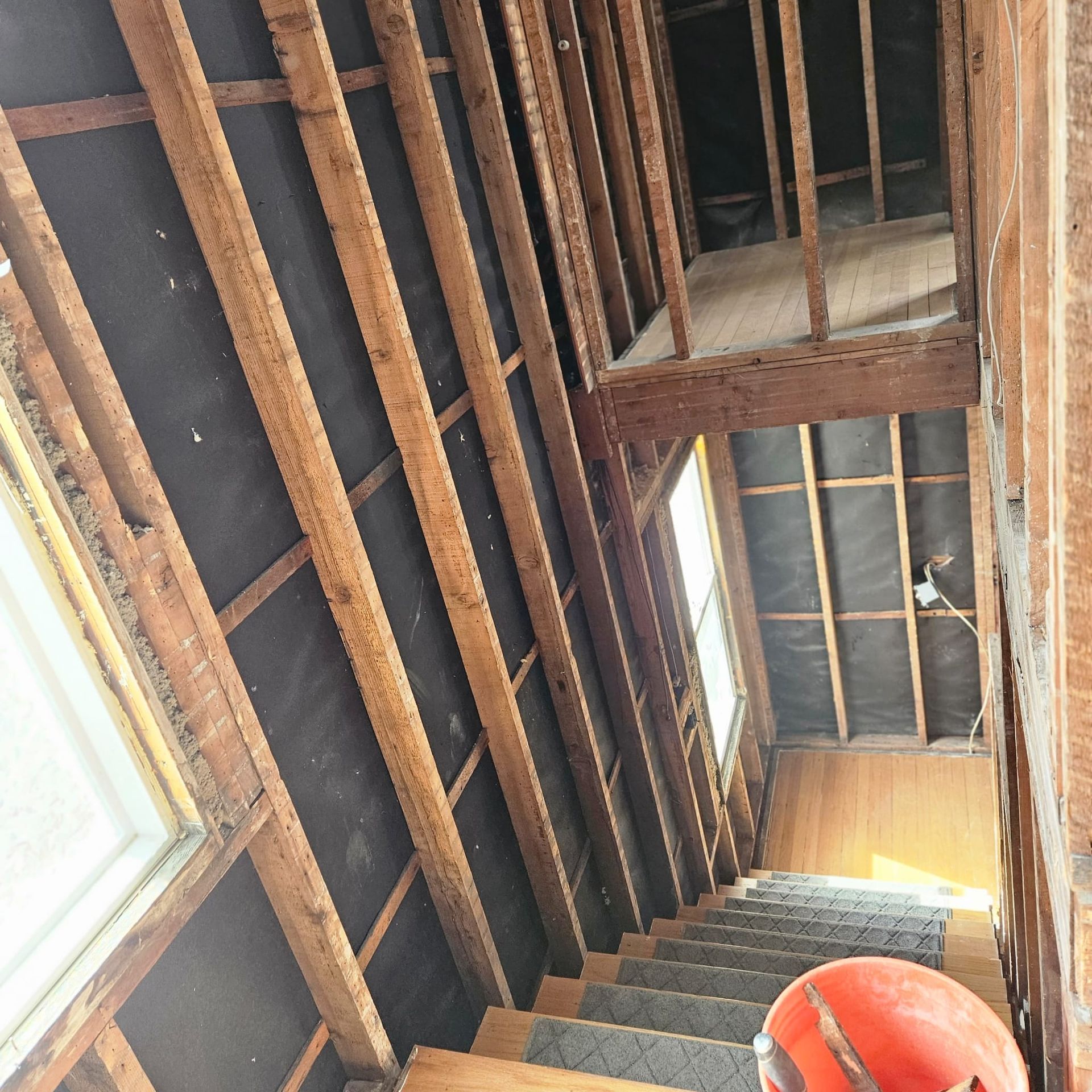 Staircase undergoing renovation; exposed wooden framing, black insulation, natural light from windows, orange hard hat visible.