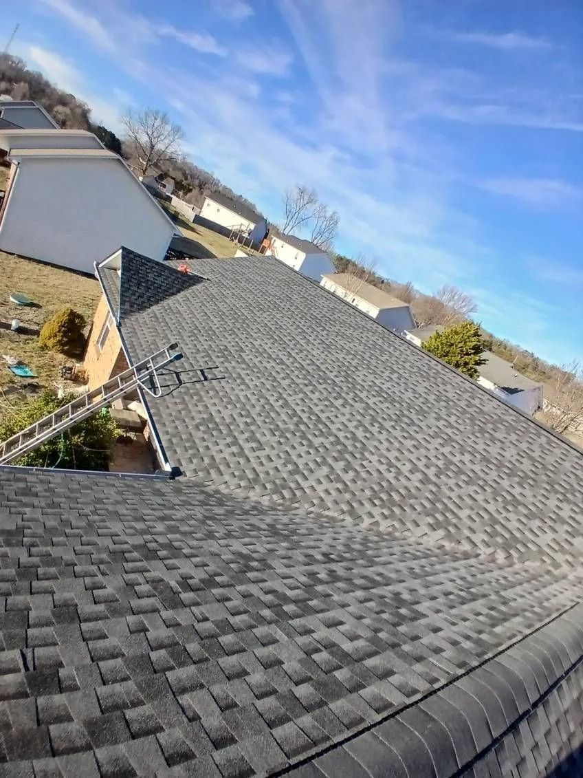 A high-angle view of a gray shingled residential roof on a sunny day, with a ladder leaning against the side.