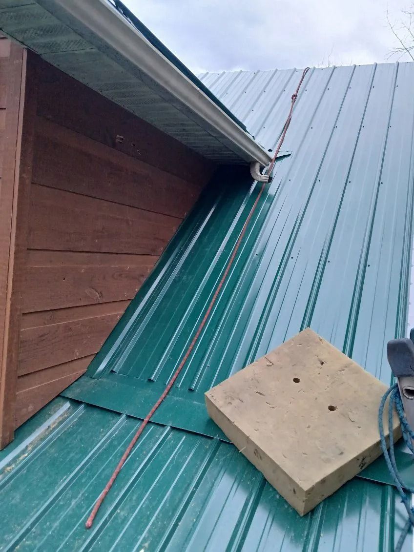 A view of a green metal roof with a wood-sided dormer, a drainage gutter, and a square wooden block resting on the slope.