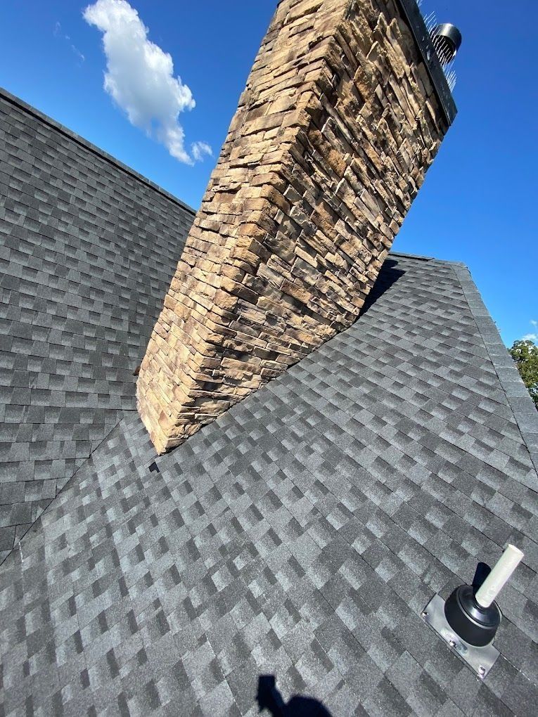 A stone chimney rises through a dark shingled roof under a bright blue sky, with a roof vent visible nearby.