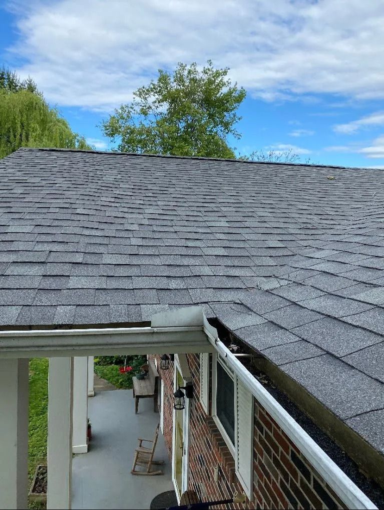 A partial view of a gray shingled roof with a damaged gutter section above a covered brick porch.