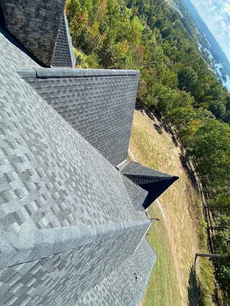 High-angle view of a grey shingled roof with multiple peaks overlooking a grassy hill and distant forest.