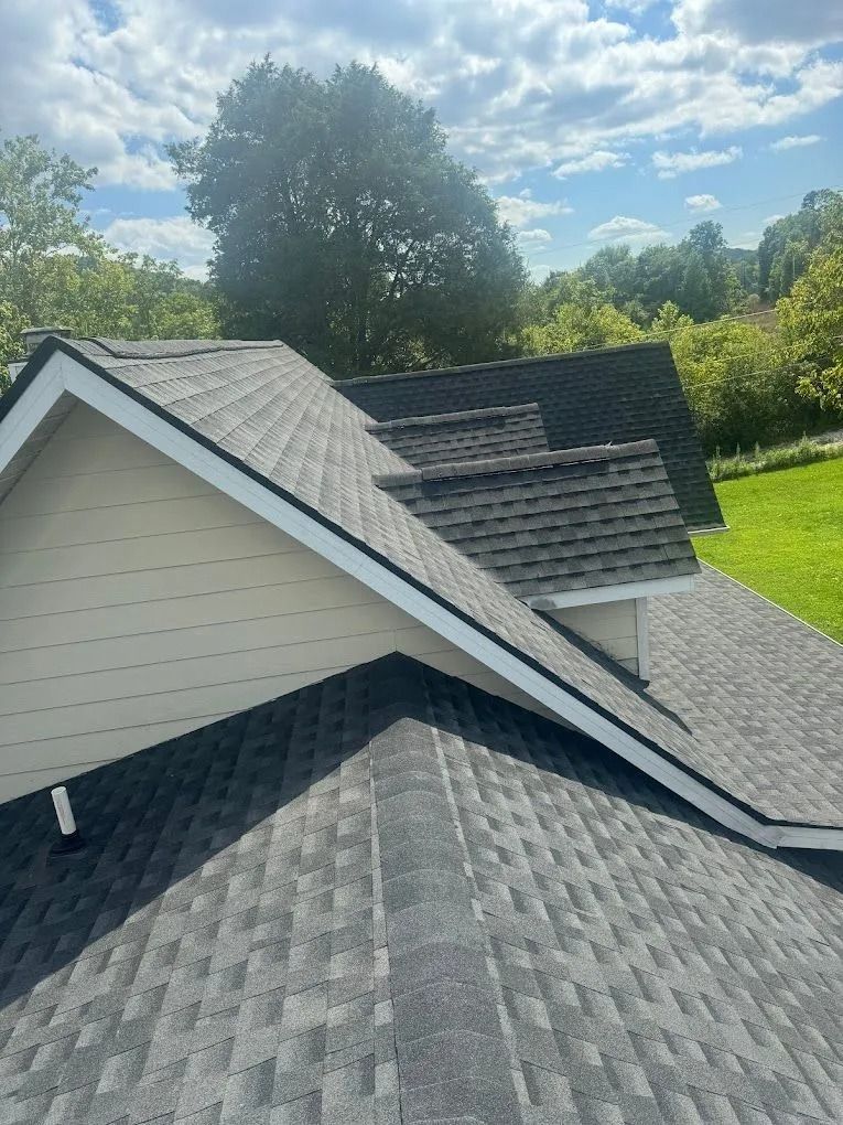 An elevated view of shingled roofs on a residential home with white trim, set against a backdrop of trees and a blue sky.